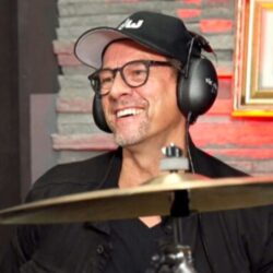 Image of Thomas Lang with a headset on sitting at a drum set with a symbol in the foreground.